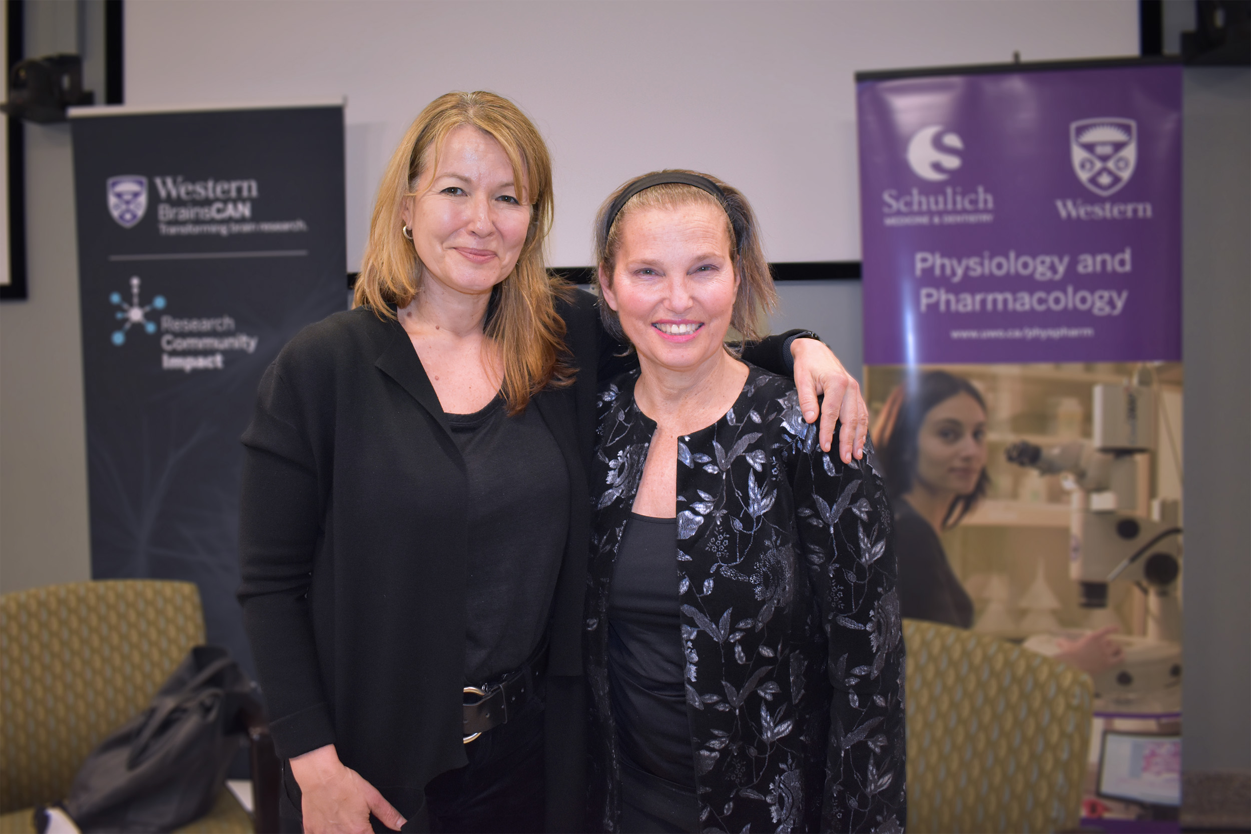 Lisa Saksida and MP Kirsty Duncan smiling at the camera with Western BrainsCAN and Schulich Physiology and Pharmacology banners in the background.